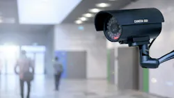 Close-up of security camera on wall with blurred background of people walking in hallway.