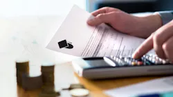 Person using calculator holding piece of paper with college cap icon and stacks of coins on table in foreground.