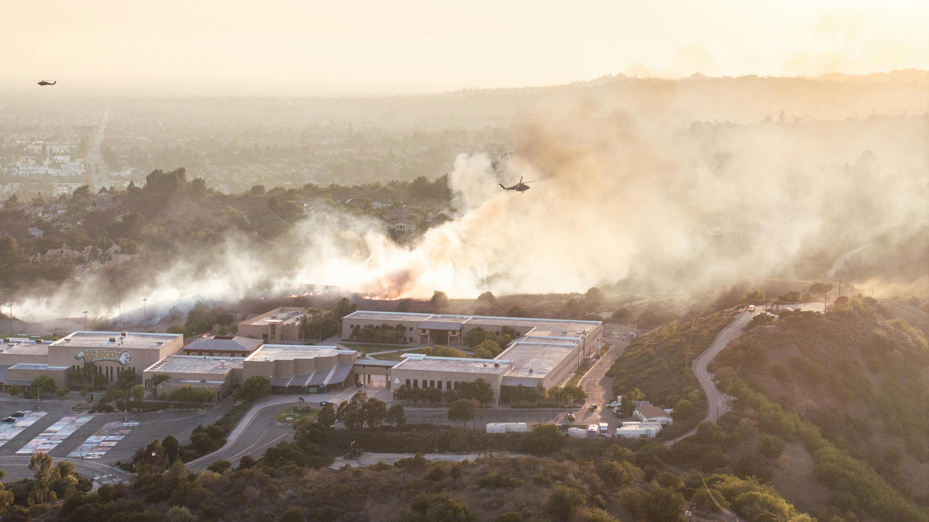 Wildfire smoke near school buildings in California with helicopters hovering overhead.