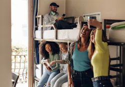 Students in dorm room with girls posing for selfie while other study in background on bunkbed.