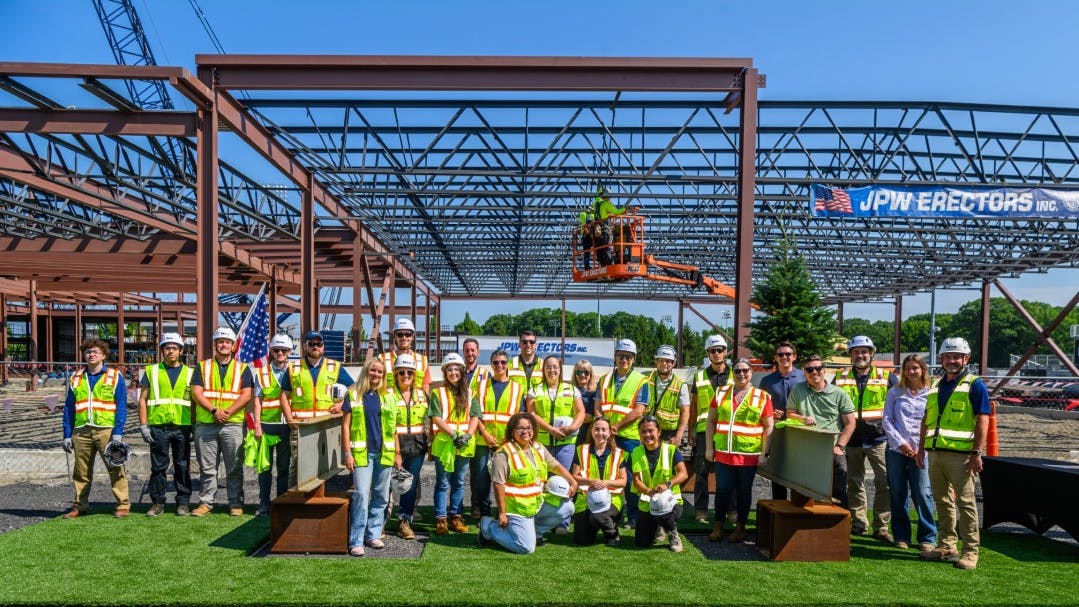 A large construction crew in safety vests pose for photo in front of steel-framed building construction project.