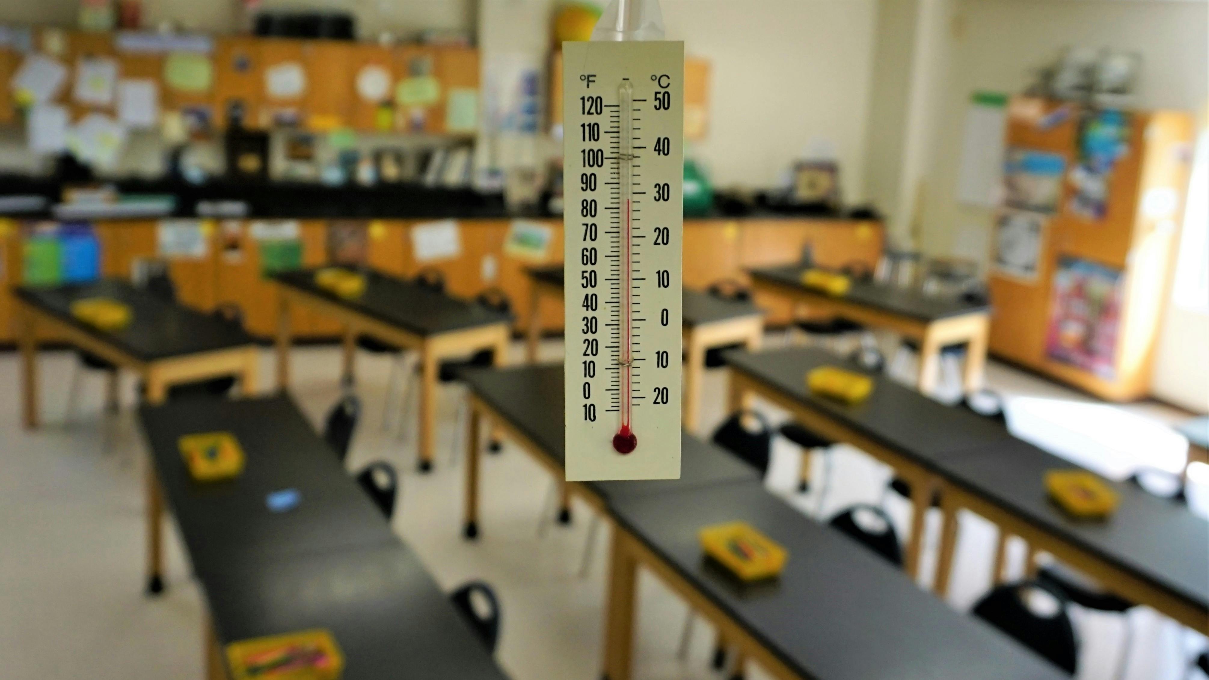 Thermometer hangs from classroom ceiling showing high temperature with desks in rows in the background.