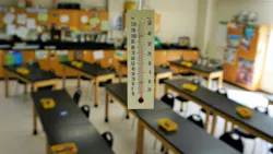 Thermometer hangs from classroom ceiling showing high temperature with desks in rows in the background.