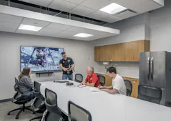 Conference room with people seating and one person standing holding equipment with large display screen on back wall.