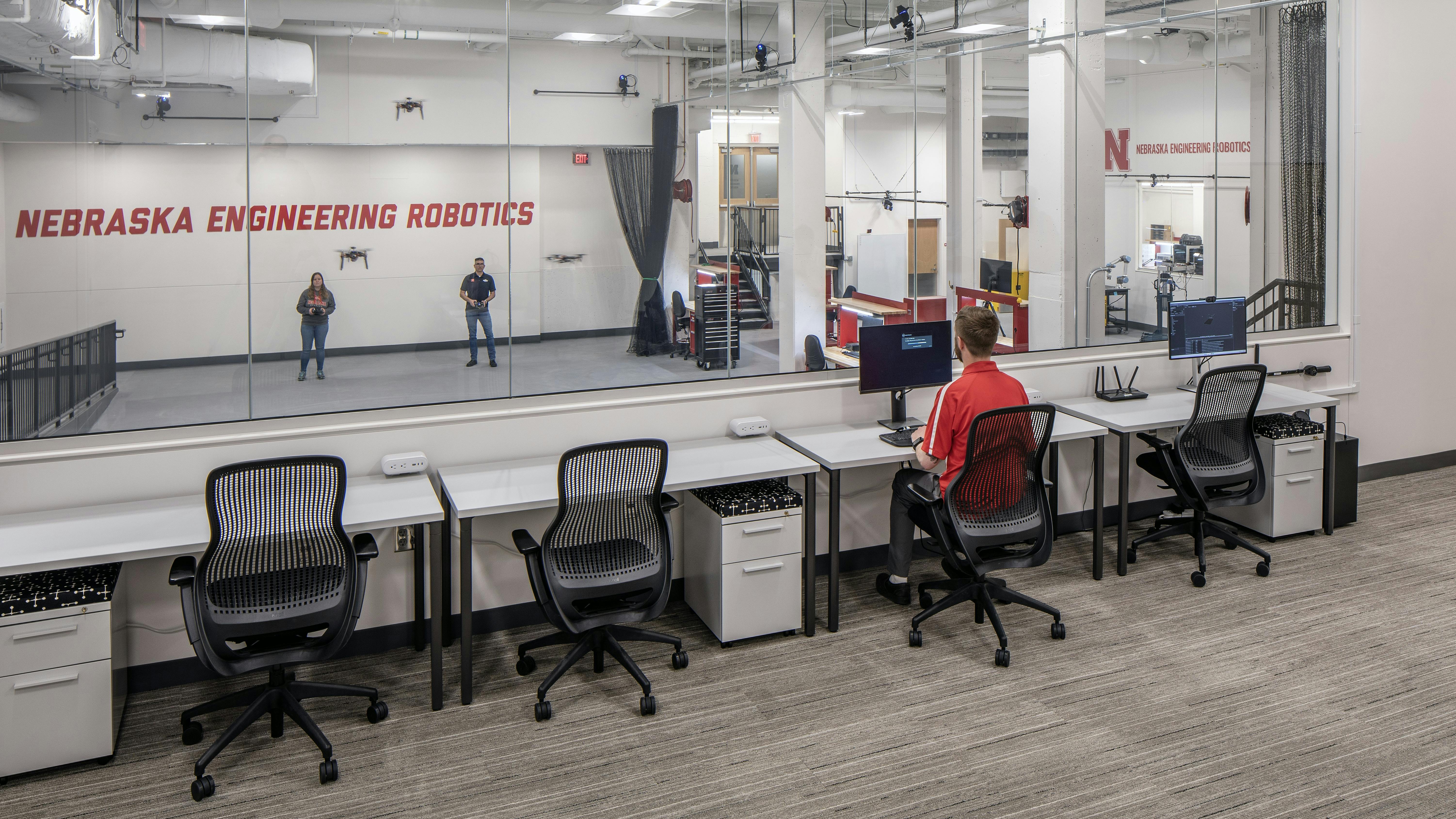 Inside observation room with glass wall where people are testing drones in other room.