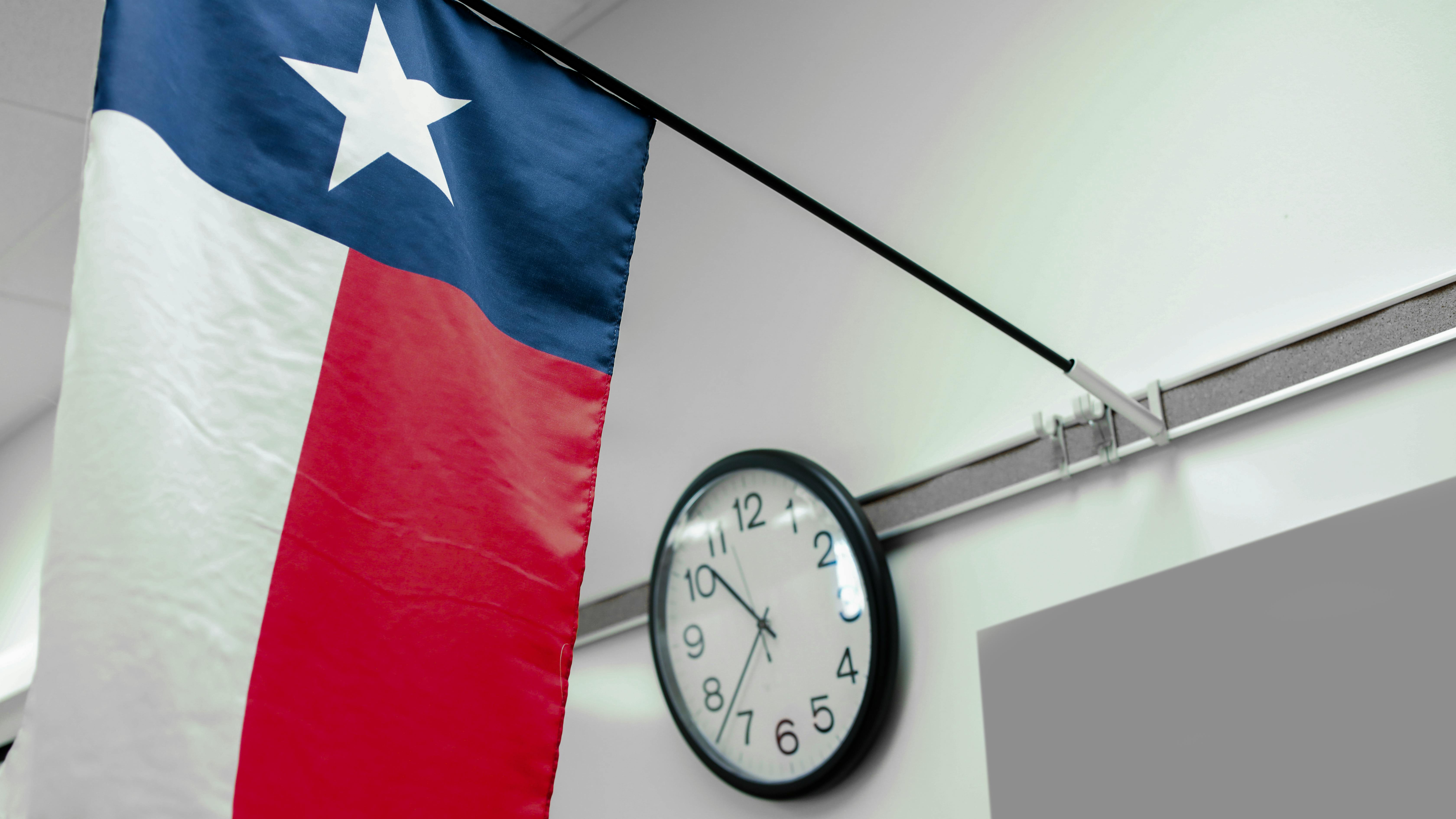 Flag of Texas hangs in front of chalkboard with wall clock in background.
