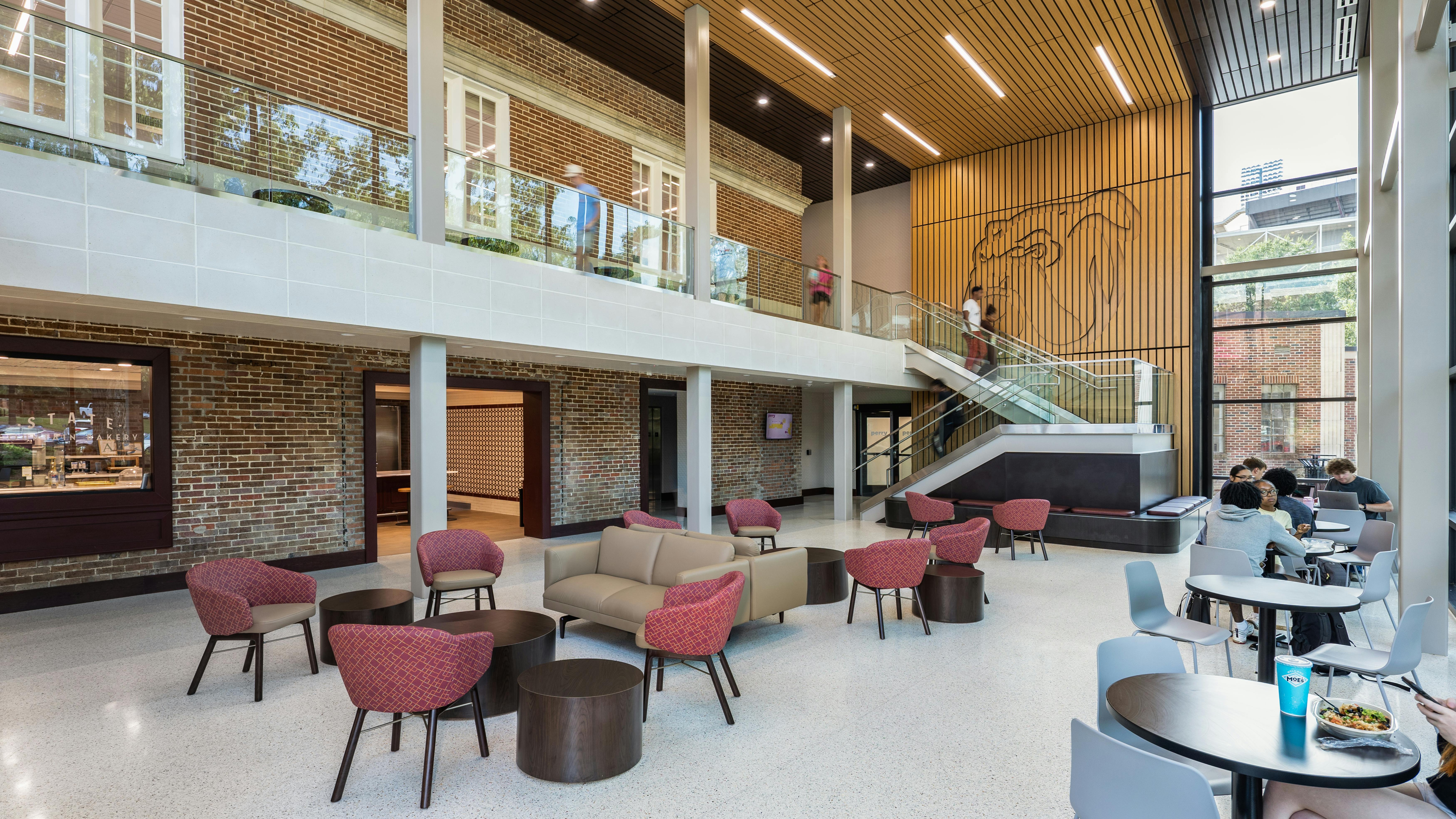 Two-story renovated dining hall with seating at bottom of staircase featuring brick and wood detailing on walls.