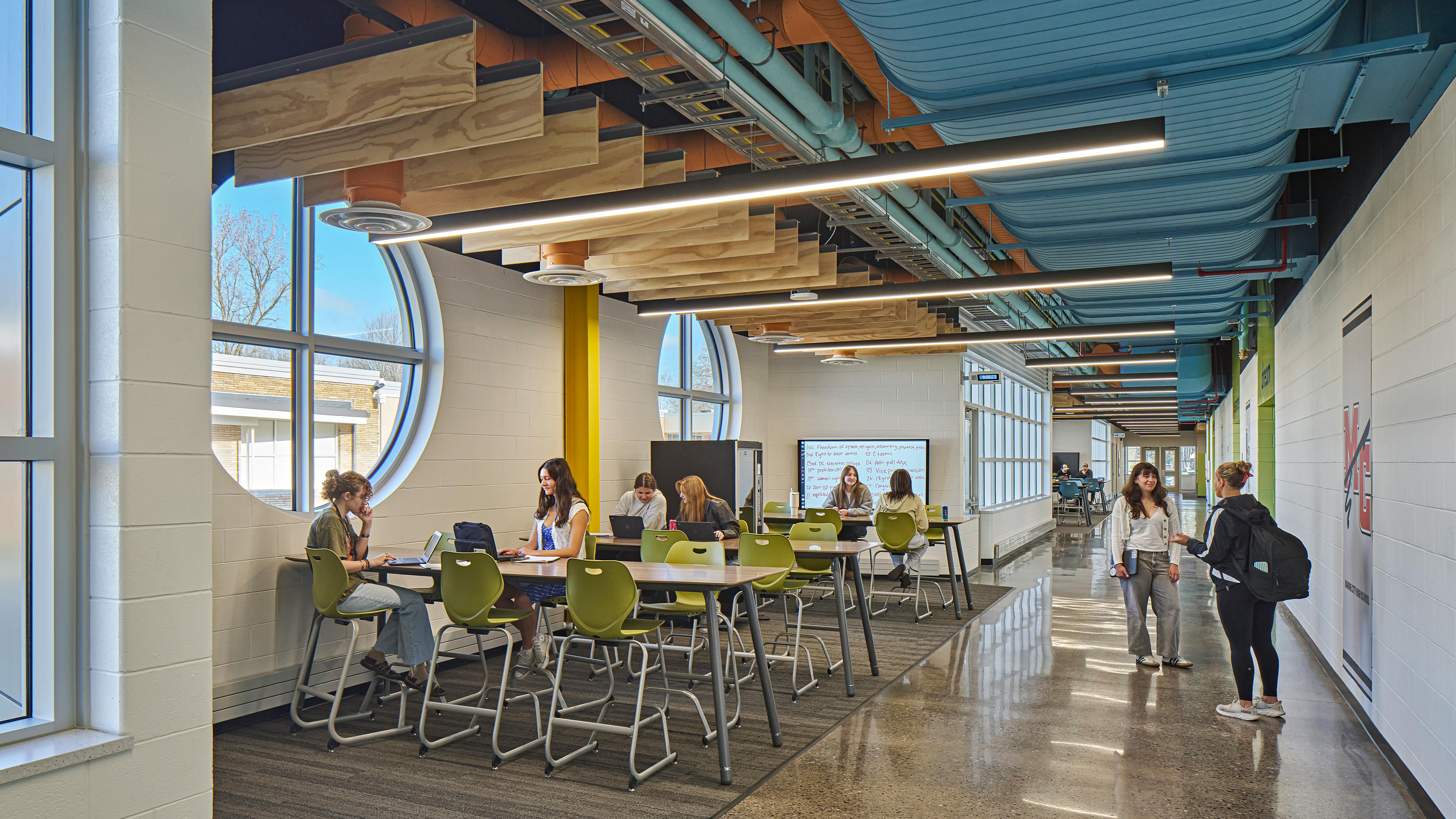 Students gather in a casual study space next to circular windows with wood ceiling baffles hanging above.