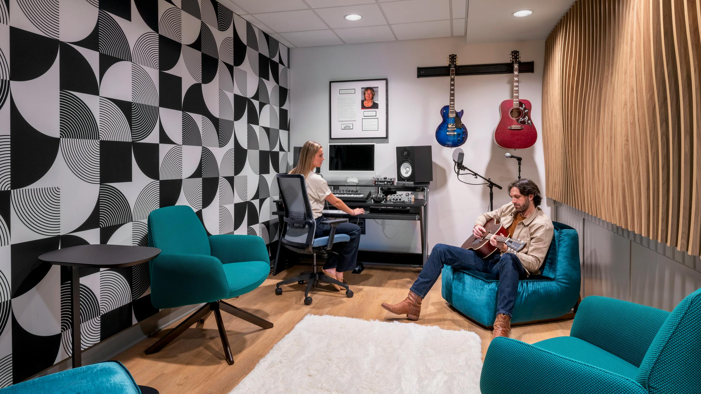 Modern music recording space with wood flooring, colorful lounge seating, geometric wallpaper and acoustic wall panels, and a musician and sound engineer seated.