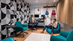 Modern music recording space with wood flooring, colorful lounge seating, geometric wallpaper and acoustic wall panels, and a musician and sound engineer seated.