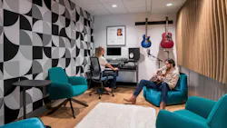 Modern music recording space with wood flooring, colorful lounge seating, geometric wallpaper and acoustic wall panels, and a musician and sound engineer seated.