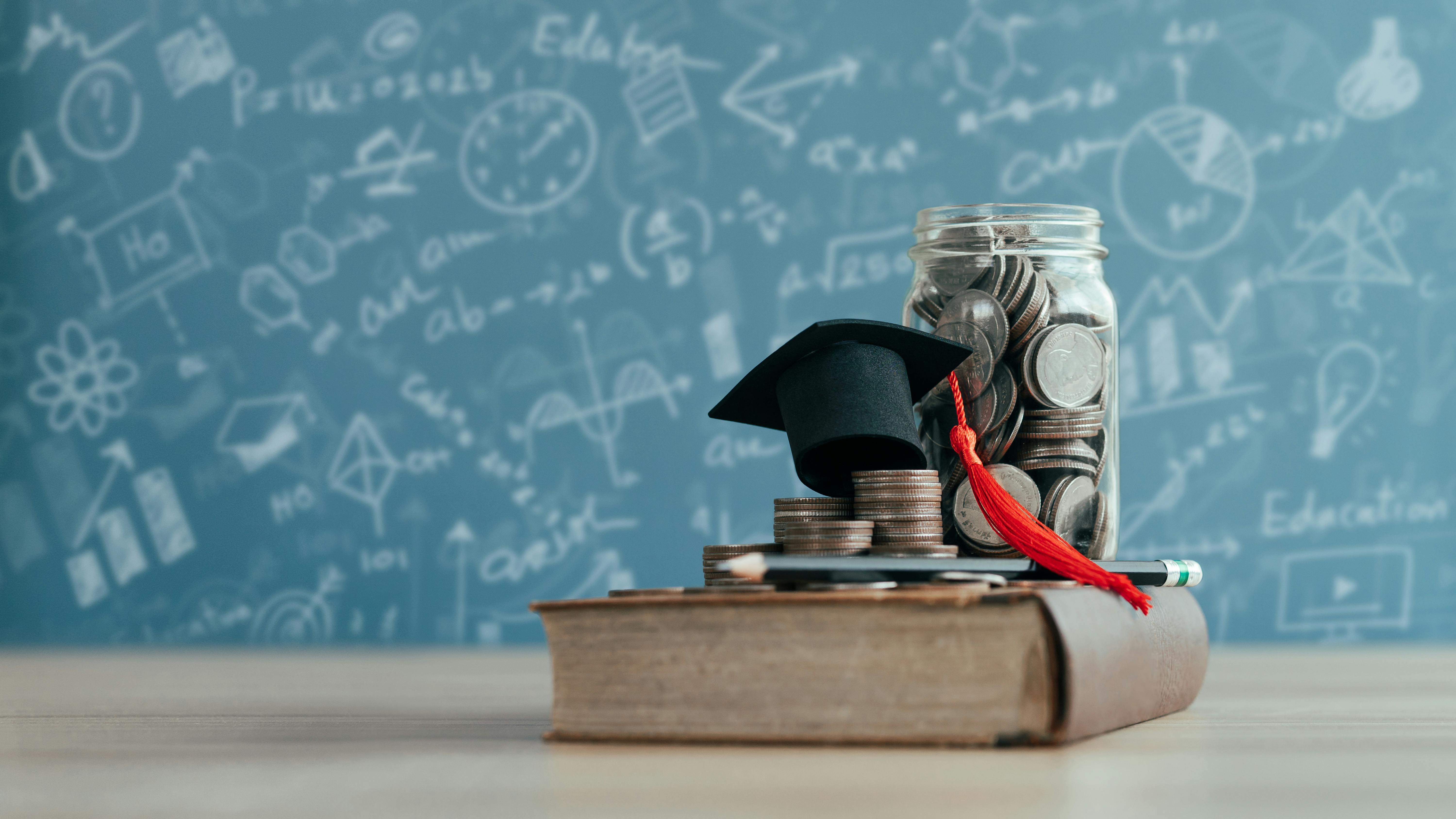 Money jar with graduation cap on top of book with blurred chalkboard in background with scientific formulas depicting education financing concept.