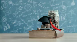 Money jar with graduation cap on top of book with blurred chalkboard in background with scientific formulas depicting education financing concept.