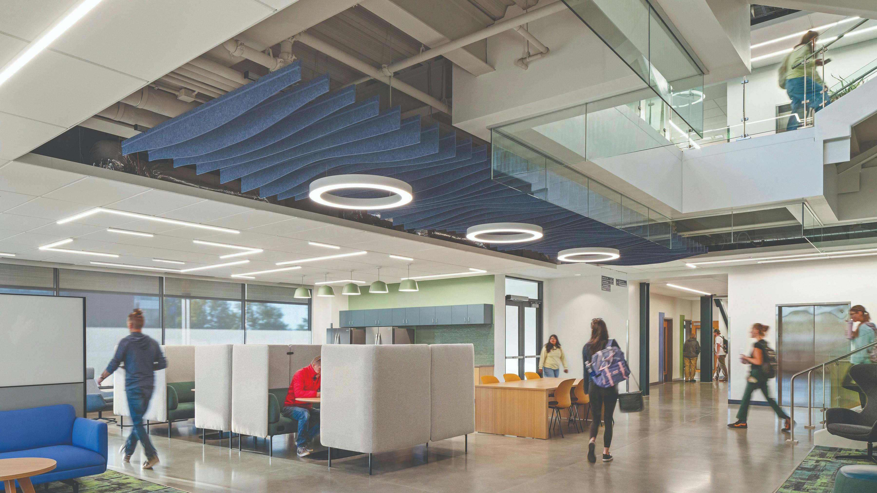Two-story common area with private booths for study beneath circular lighting fixtures and acoustic baffles near staircase with students descending.