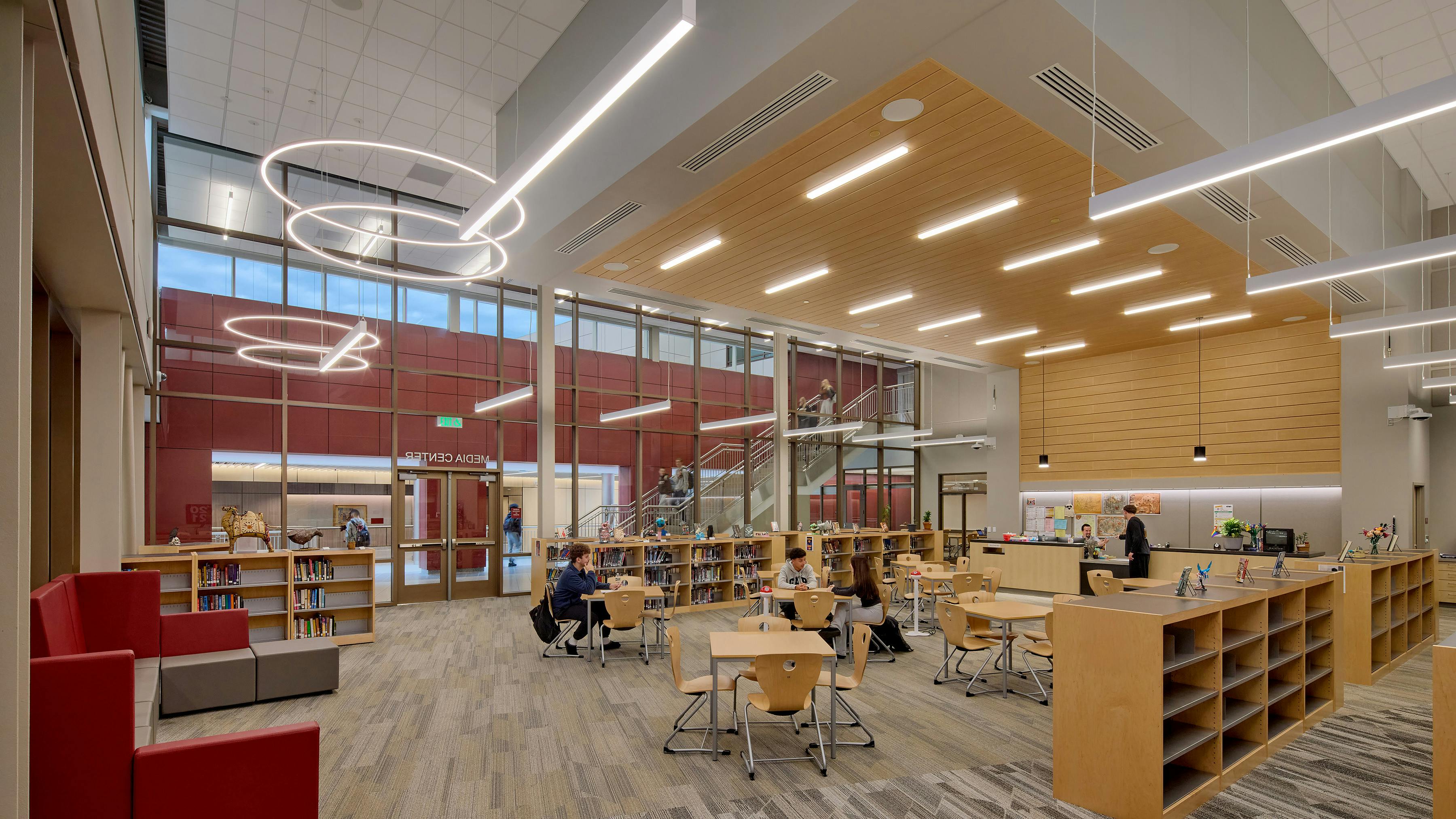 Large, open media center with bookshelves and students sitting at desks under circular and linear lighting fixtures.