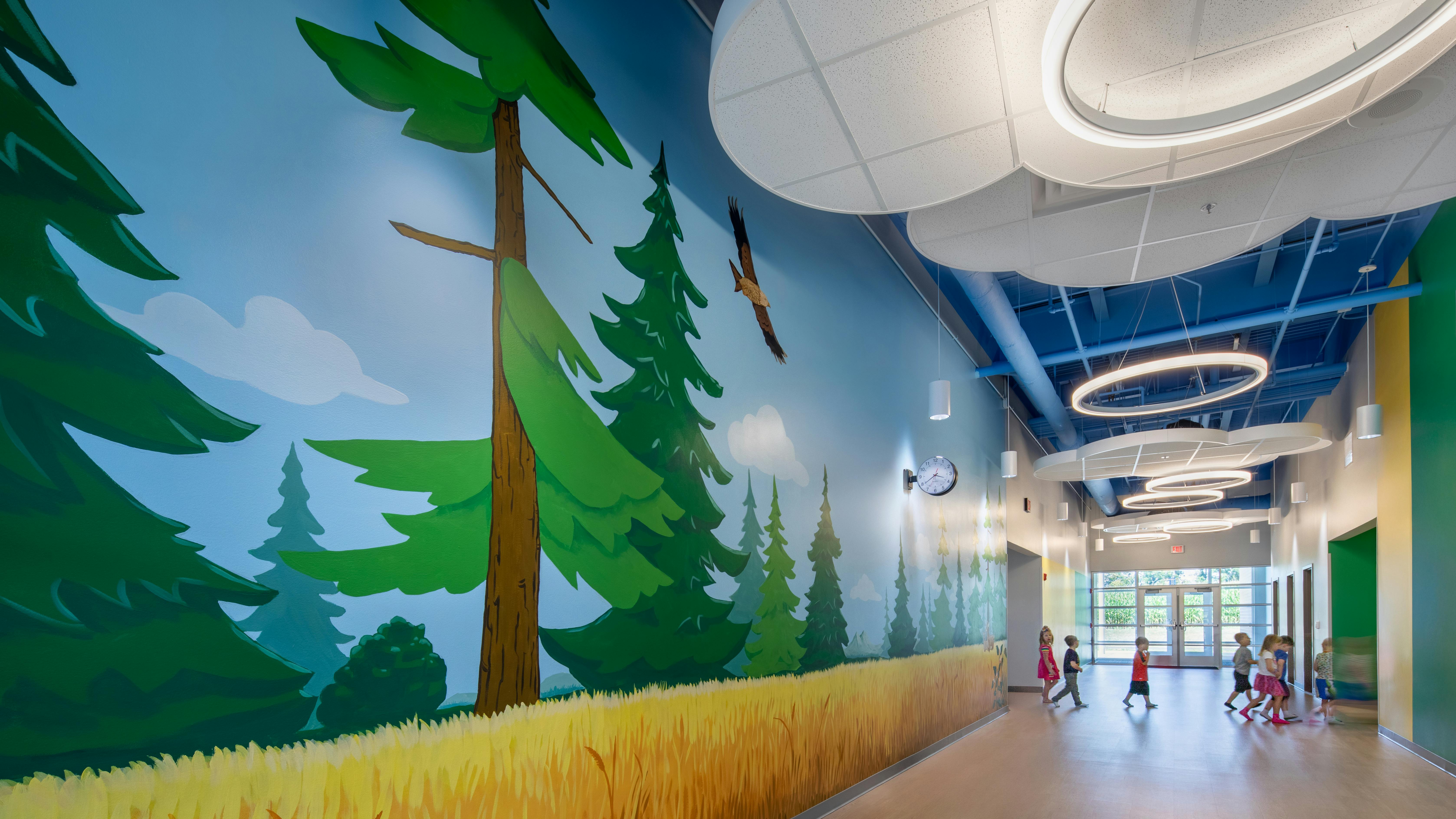 Large, colorful forest-themed wall mural in hallway of pre-K school with circular light fixtures above and children walking in distance.