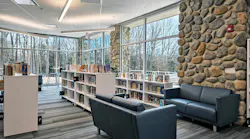 Modern library/media center with lounge seating next to natural stone wall in foreground with bookshelves near floor-to-ceiling windows in background with linear lighting fixtures above.