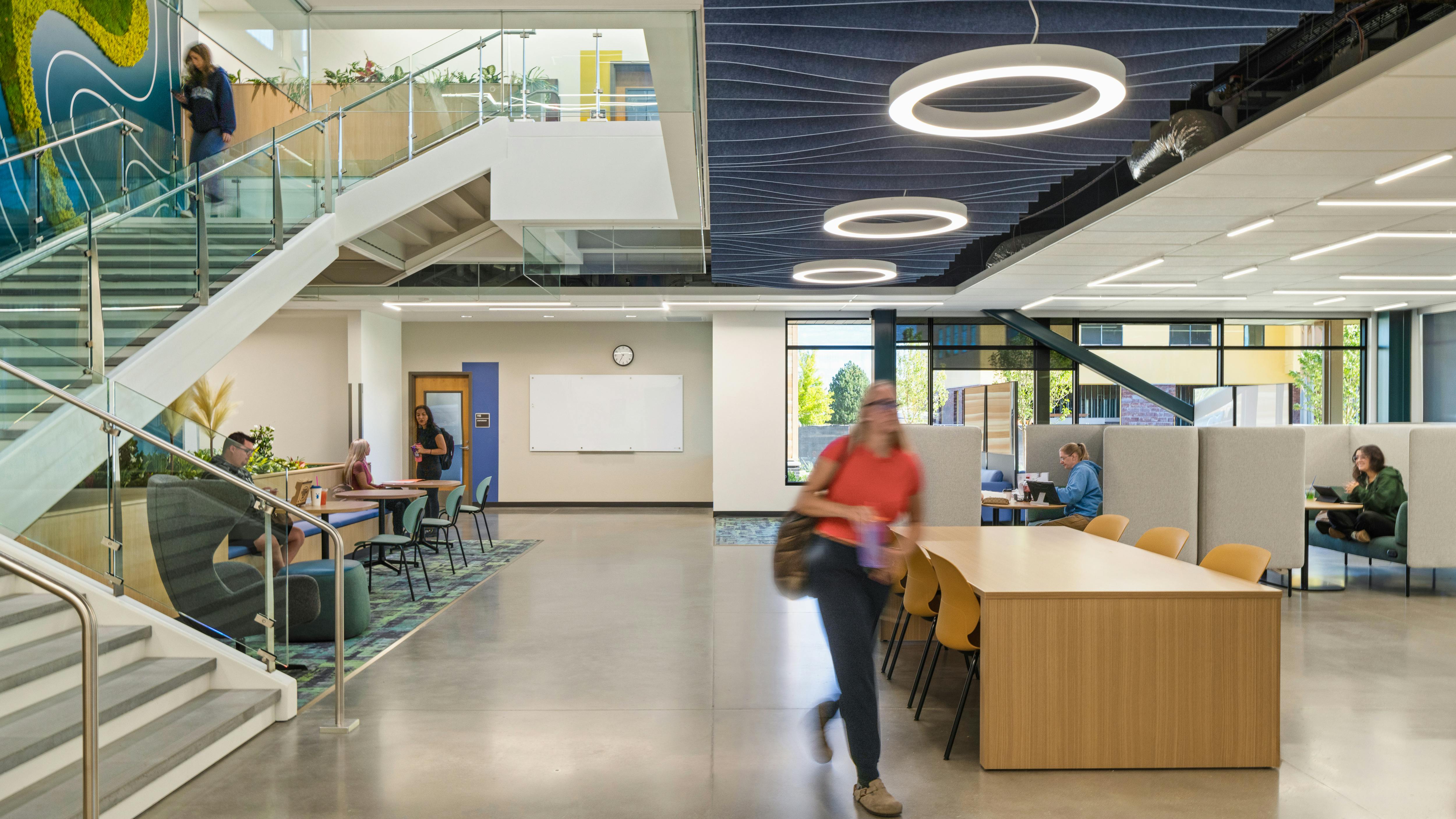 Two-story learning facility with staircase and people sitting at various seating and study areas. Acoustic baffles and circular lighting fixtures at ceiling.
