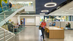 Two-story learning facility with staircase and people sitting at various seating and study areas. Acoustic baffles and circular lighting fixtures at ceiling.