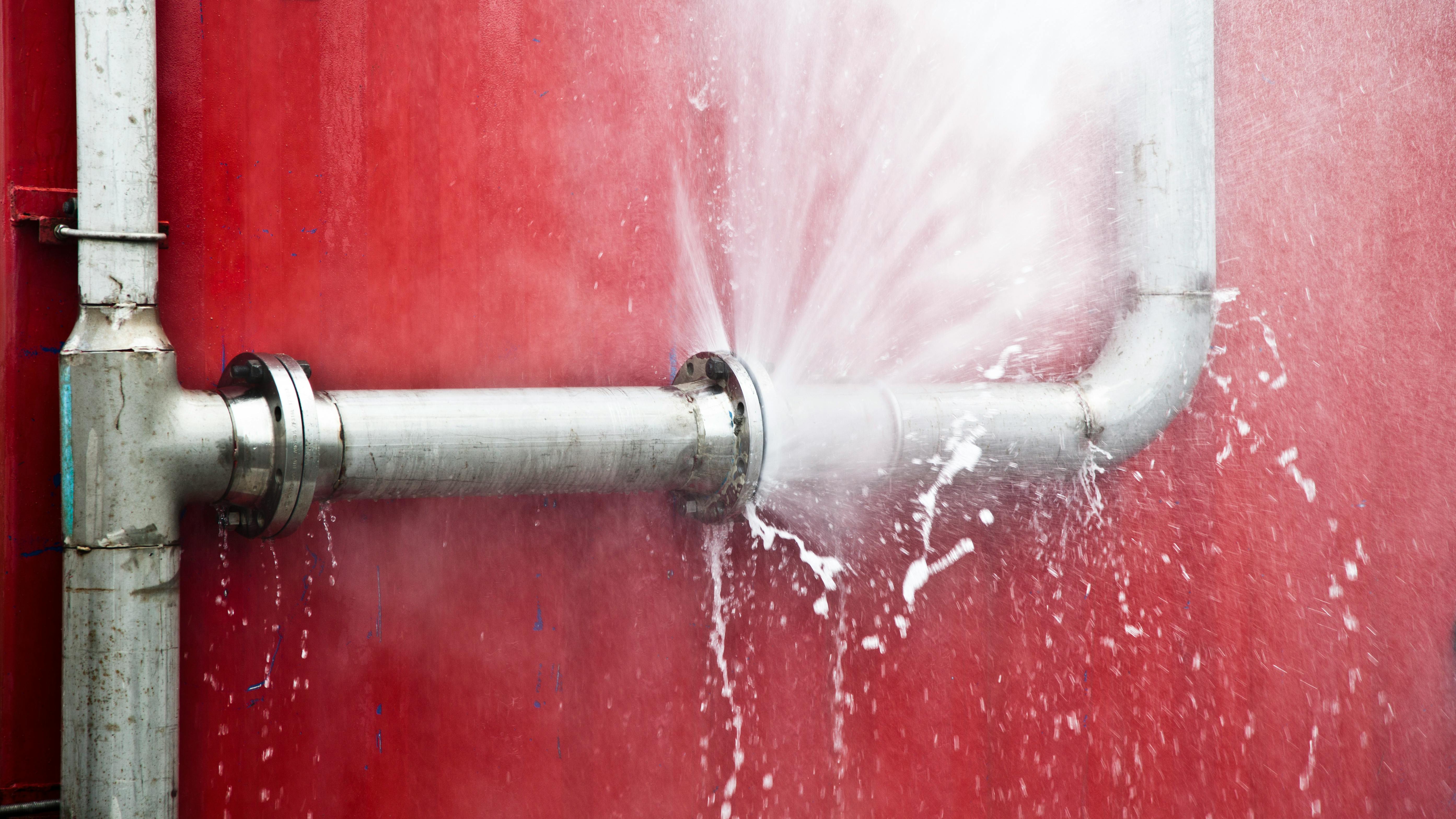 Close-up of pipe bursting with water against red wall.