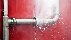 Close-up of pipe bursting with water against red wall.