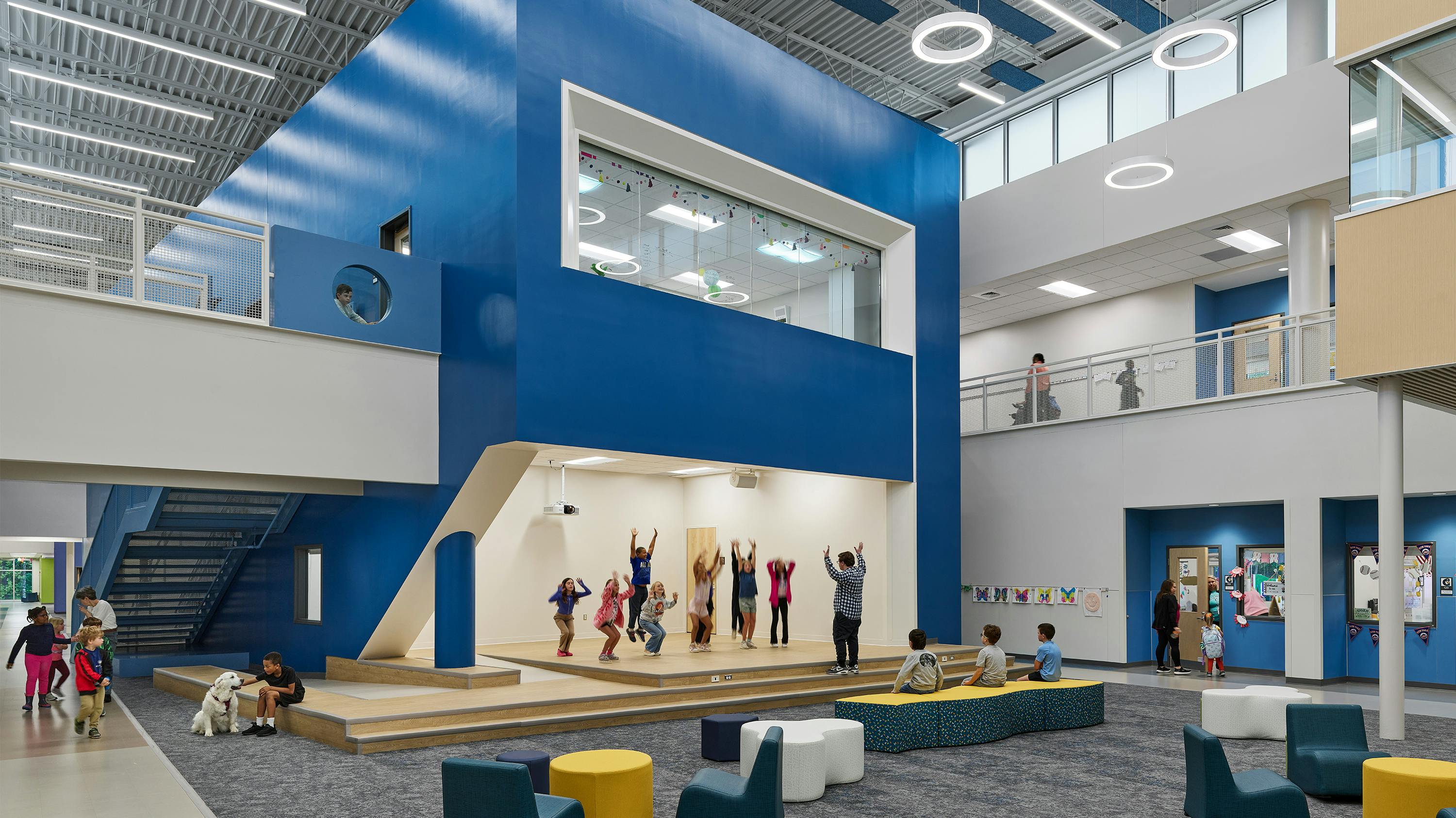 Large two-story atrium space with casual seating and a stage area with kids exercising with teacher.