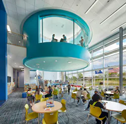 Large-two story gathering space with tables and chairs next to floor-to-ceiling windows.