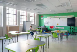 Child sits alone in daylit classroom with tables, chairs, and whiteboard with TV on the side.