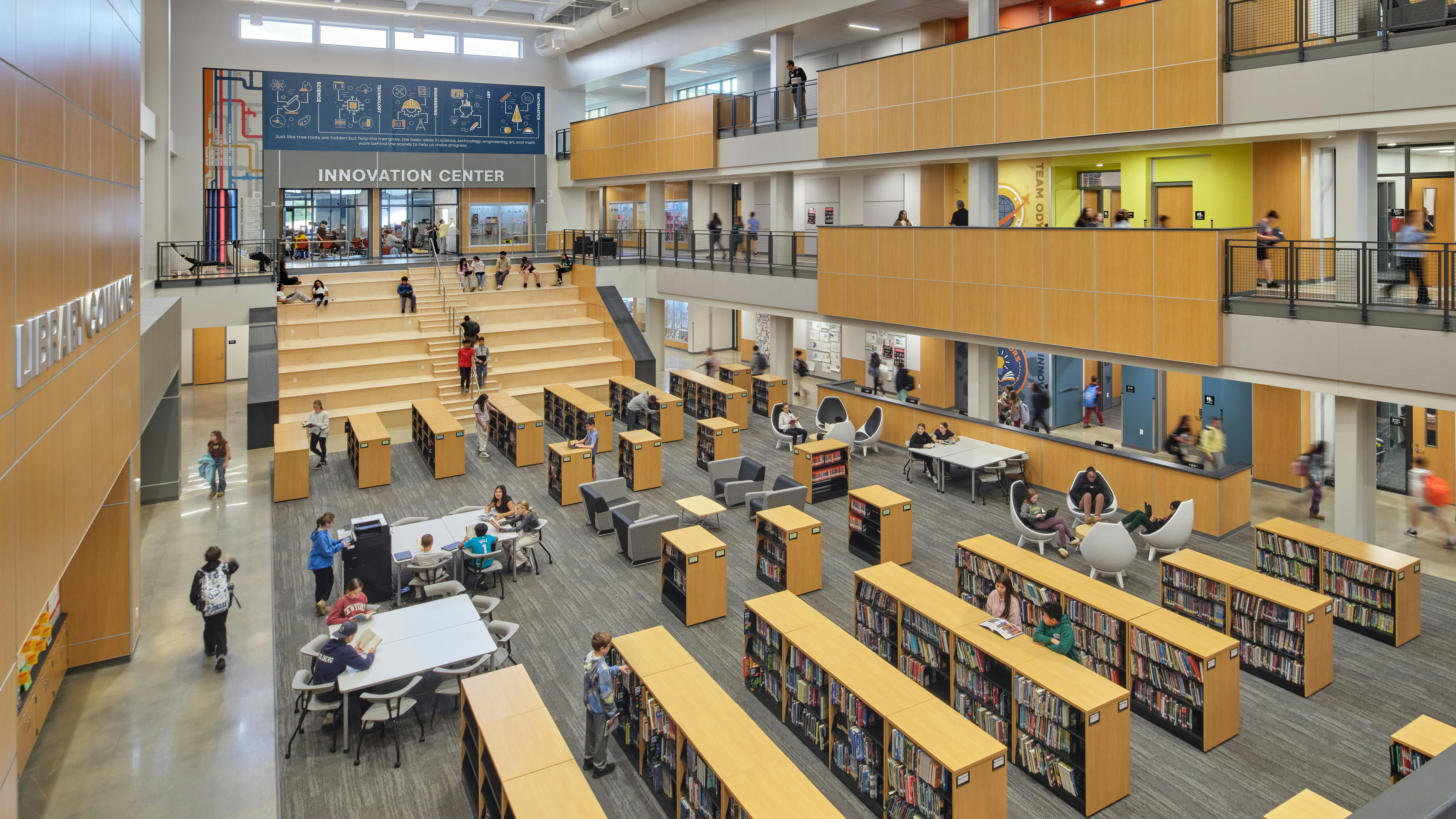 Large, open three-story learning commons with stadium seating/staircase and rows of bookshelves with seating and tables for study in between.