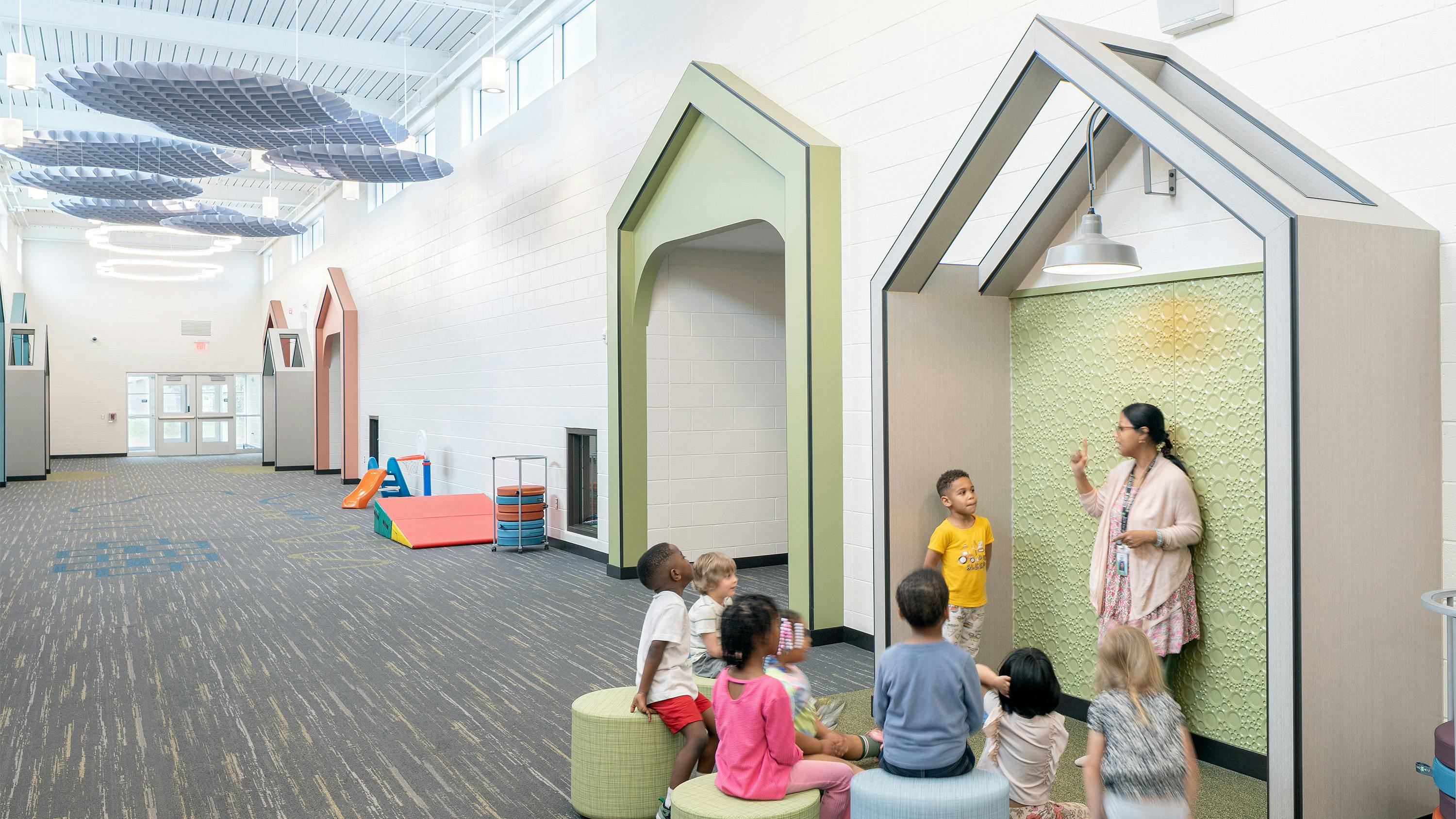 Students sit and listen to teacher standing in a geometric nook space in long hallway that features carpeting and circular lighting and waffle-shaped acoustic baffles overhead.
