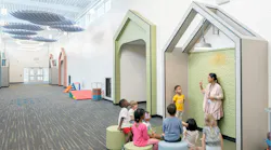 Students sit and listen to teacher standing in a geometric nook space in long hallway that features carpeting and circular lighting and waffle-shaped acoustic baffles overhead.