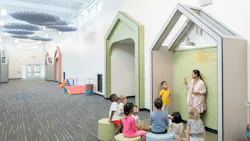 Students sit and listen to teacher standing in a geometric nook space in long hallway that features carpeting and circular lighting and waffle-shaped acoustic baffles overhead.
