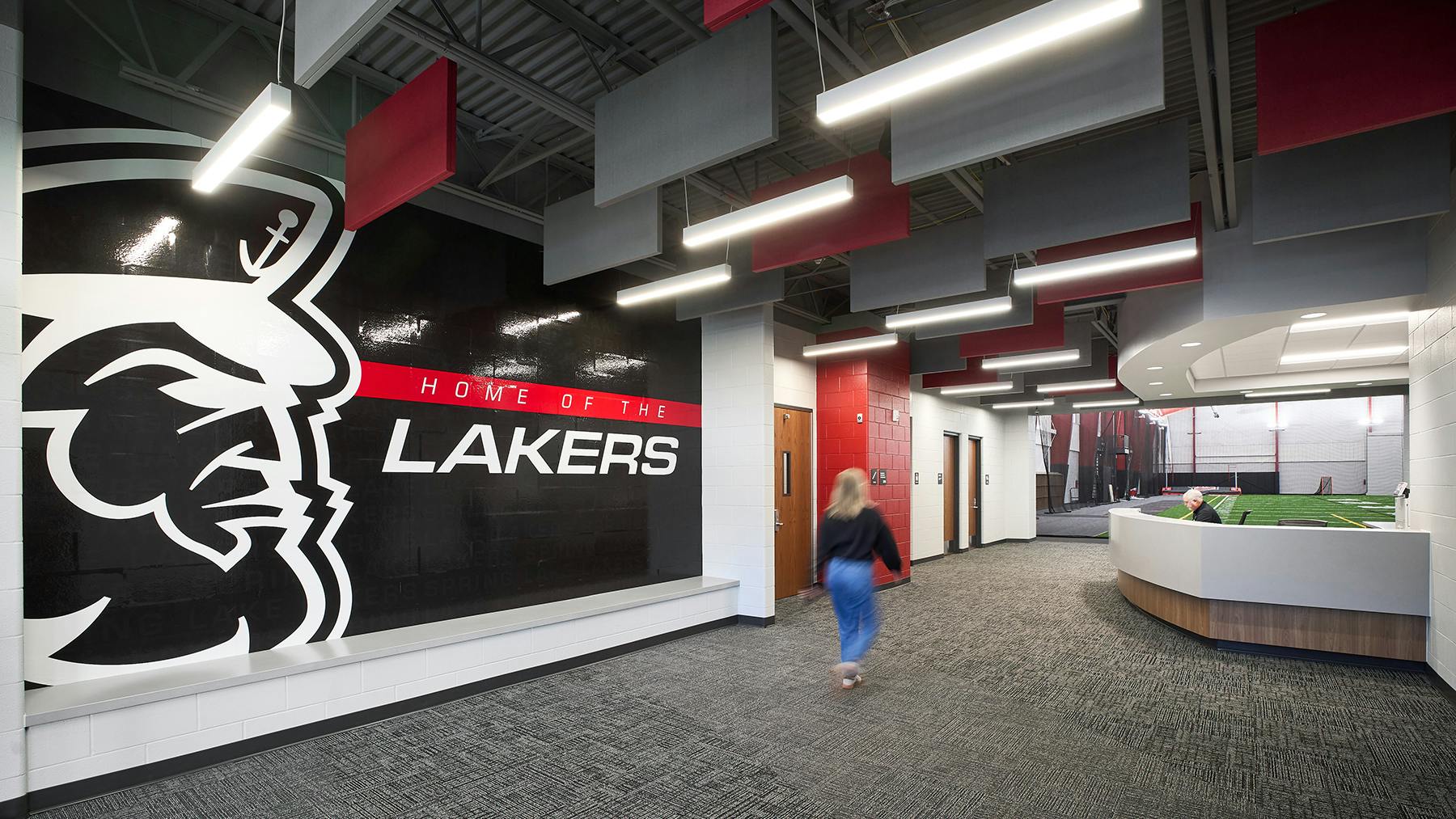 Woman walks toward reception area in hallway of new athletic faclity featuring large, branded graphic of school mascot and name with acoustic baffles and linear lighting fixtures above.