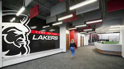 Woman walks toward reception area in hallway of new athletic faclity featuring large, branded graphic of school mascot and name with acoustic baffles and linear lighting fixtures above.