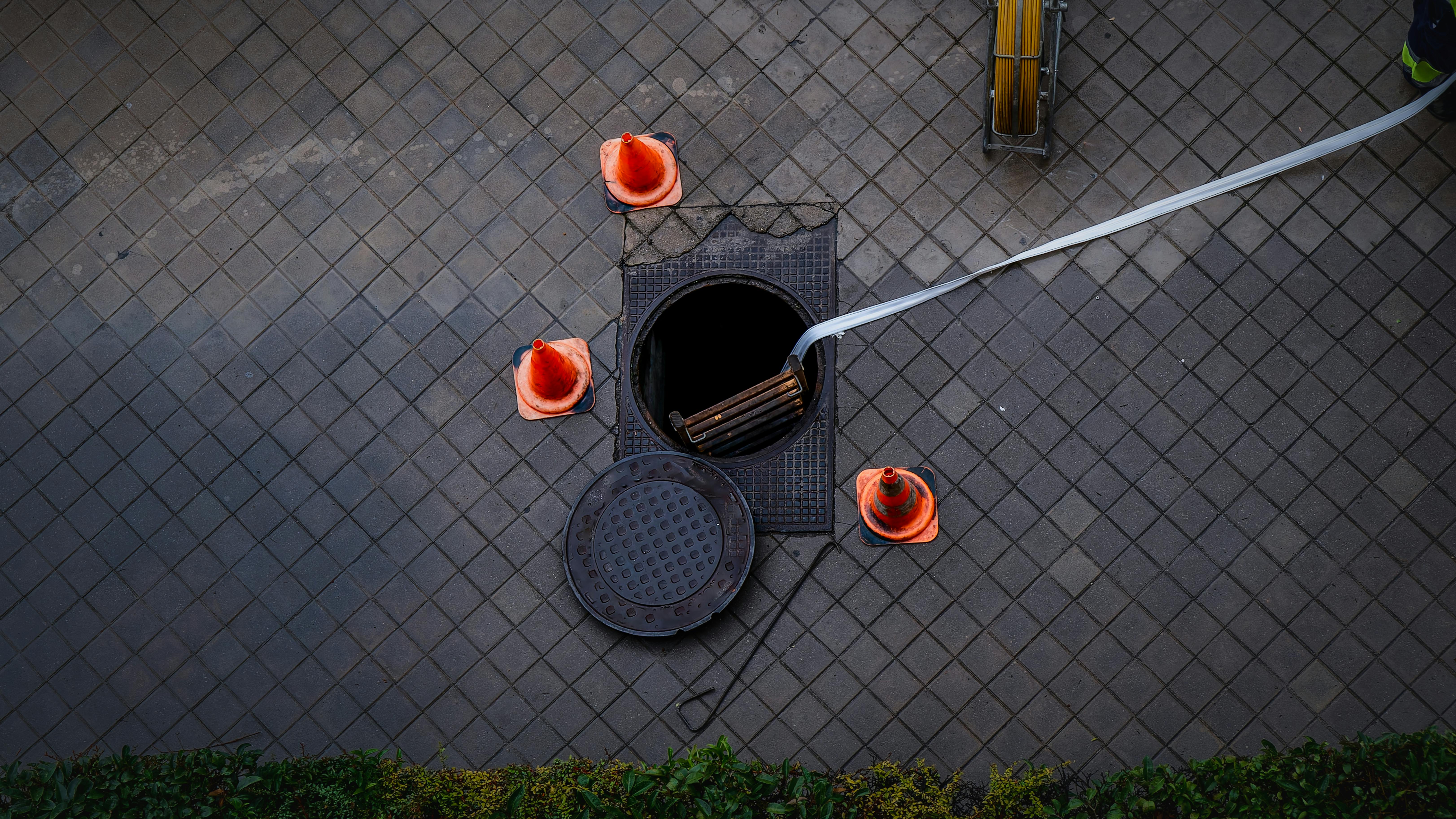 Overhead view of sewer opening on dark sidewalk with manhole cover removed and three safety cones surrounding it.