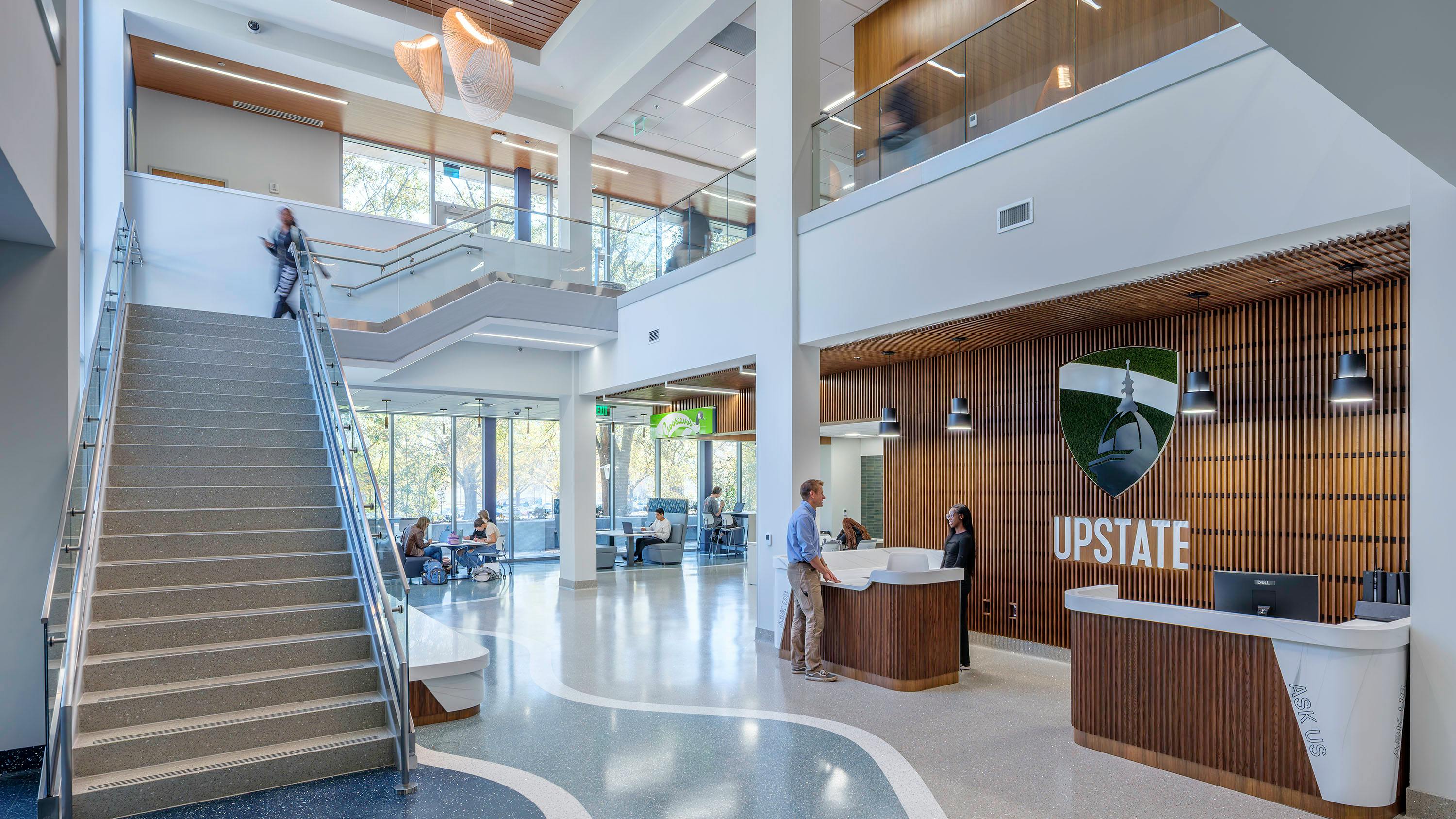 Large, two-story lobby entrance to library with reception desk featuring wood wall paneling next to a staircase with windows to outdoors in background.