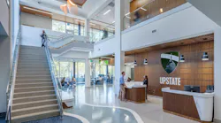 Large, two-story lobby entrance to library with reception desk featuring wood wall paneling next to a staircase with windows to outdoors in background.