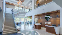 Large, two-story lobby entrance to library with reception desk featuring wood wall paneling next to a staircase with windows to outdoors in background.