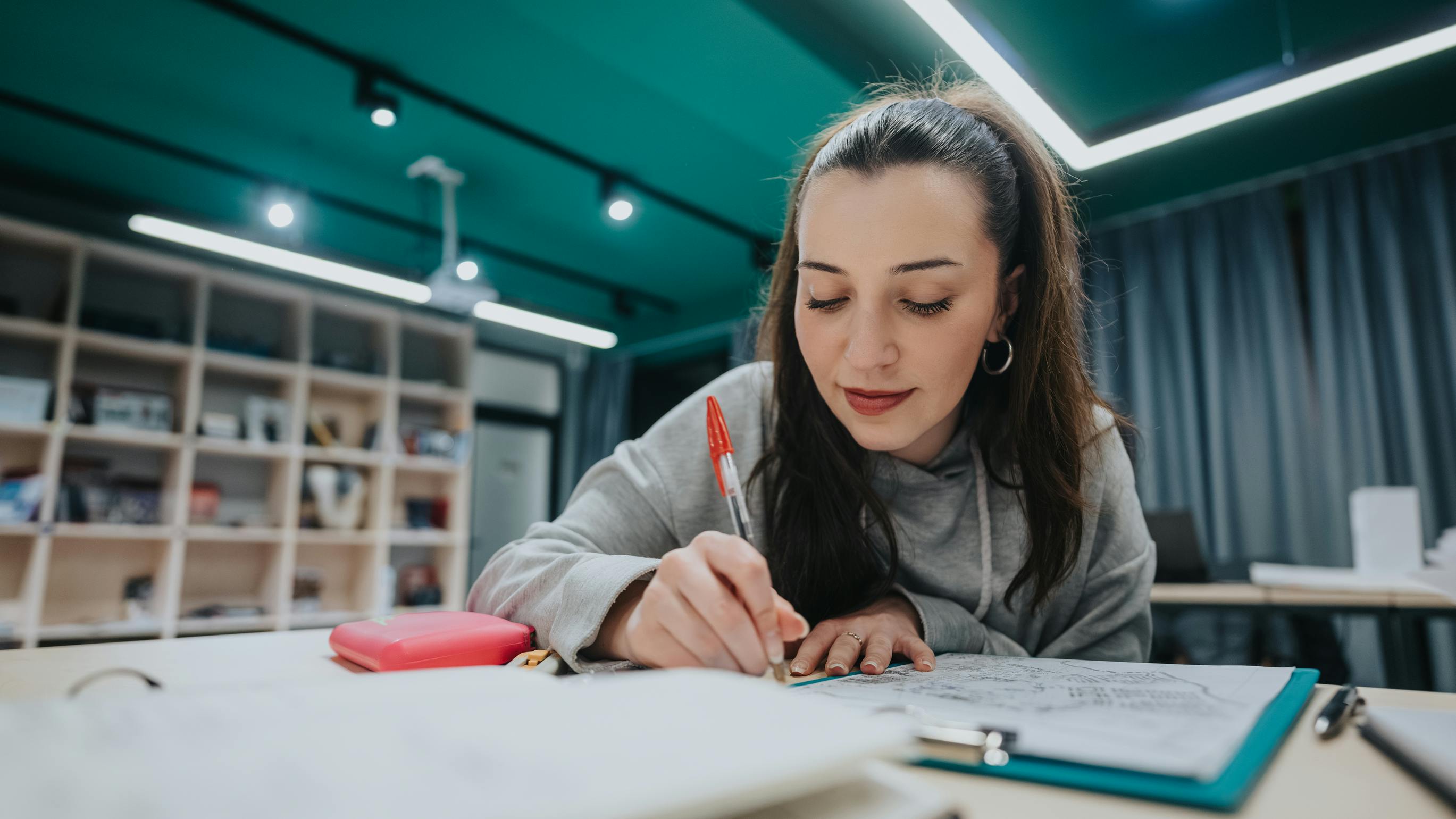 A student concentrates on sketching at a desk in a bright, modern collaborative space. Supplies include a red pencil case and papers, with shelving and soft lighting in the background.