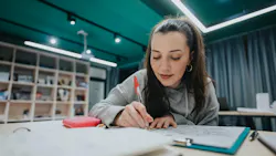 A student concentrates on sketching at a desk in a bright, modern collaborative space. Supplies include a red pencil case and papers, with shelving and soft lighting in the background.