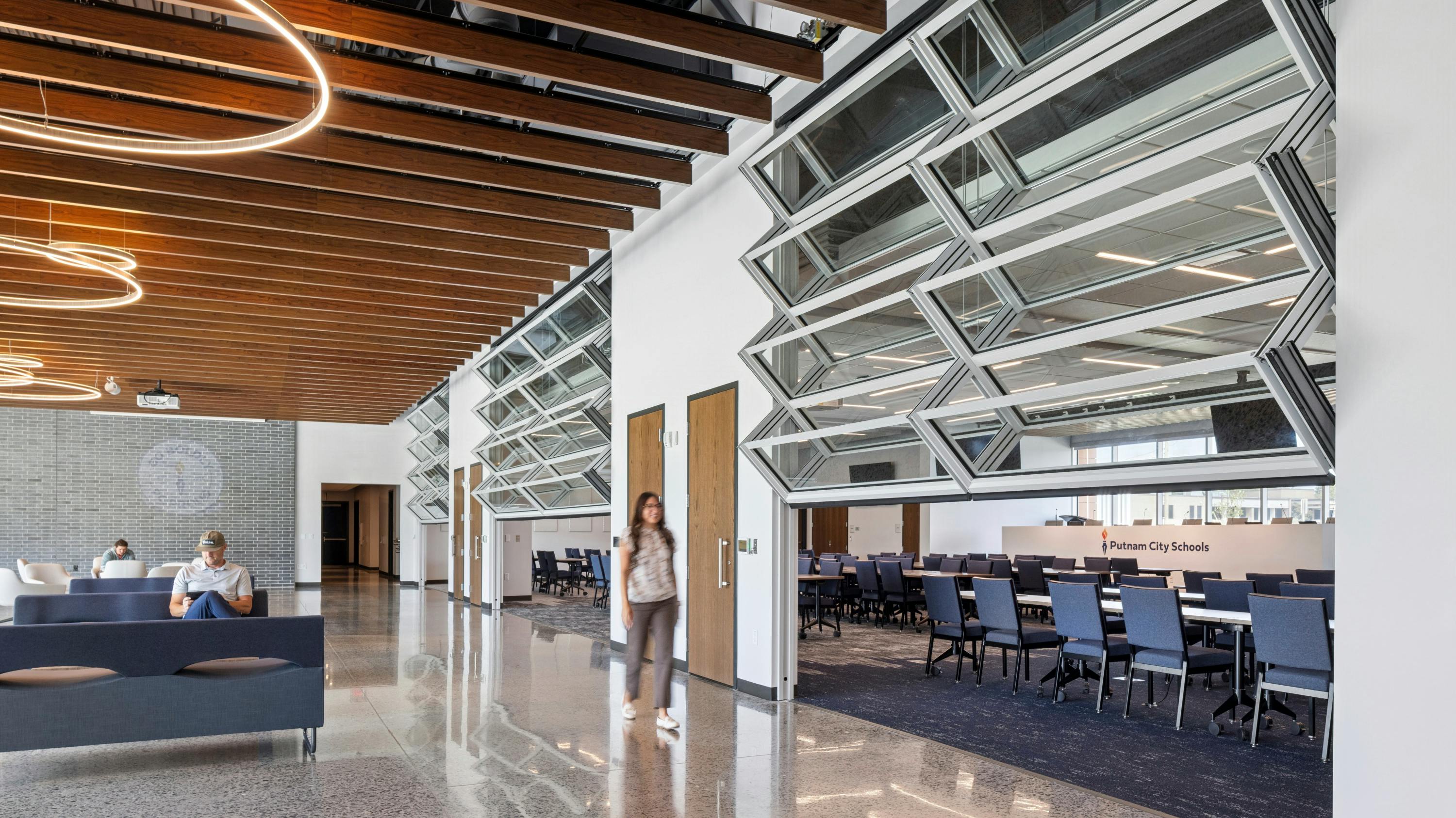 Student walks past half-opened, glass garage doors that open to a classroom with others sit in adjacent lounge area beneath wood-slatted ceilings with circular lighting fixtures.