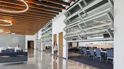 Student walks past half-opened, glass garage doors that open to a classroom with others sit in adjacent lounge area beneath wood-slatted ceilings with circular lighting fixtures.
