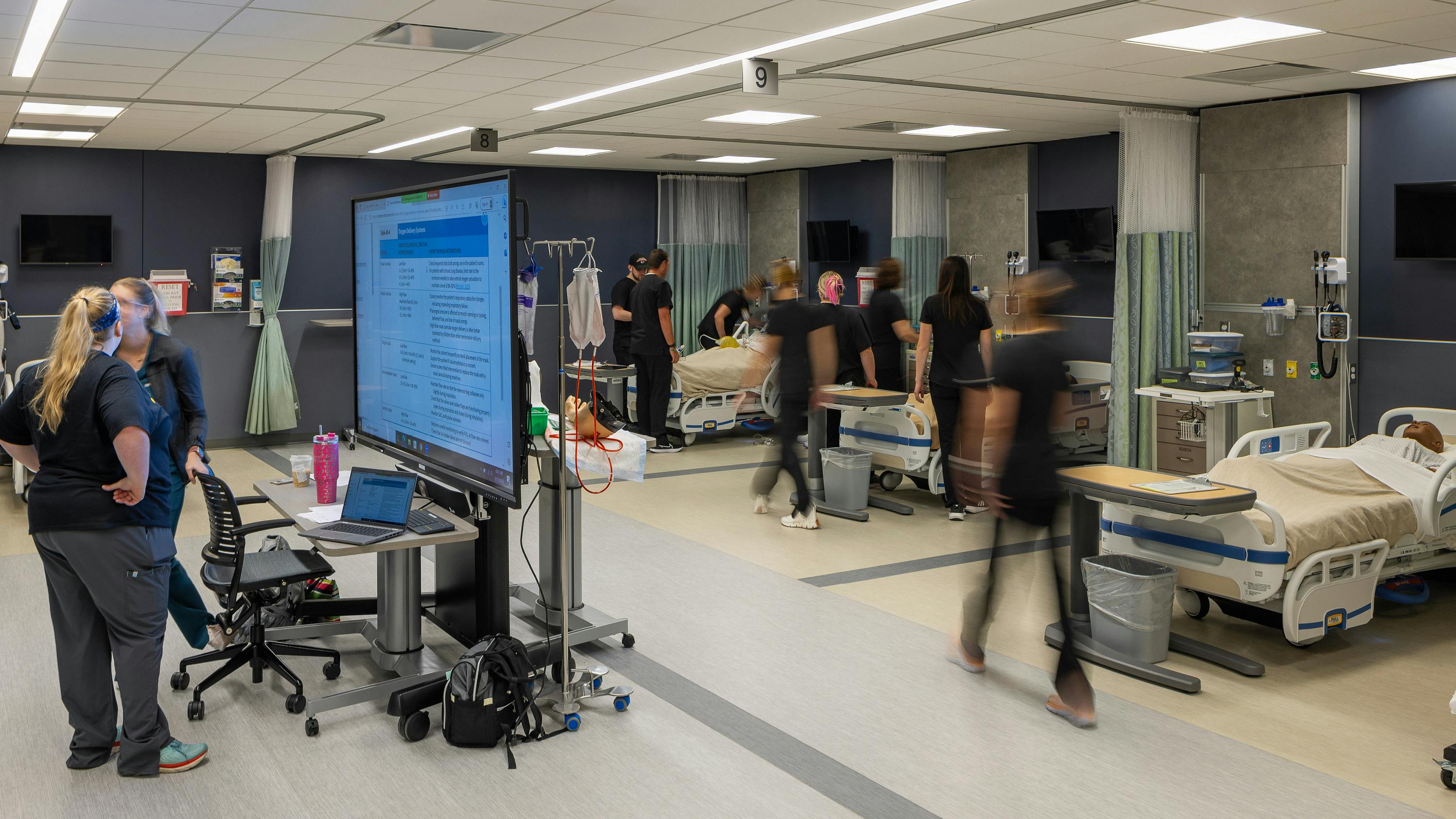 Nursing students move about a busy classroom simulation of a hospital wing.