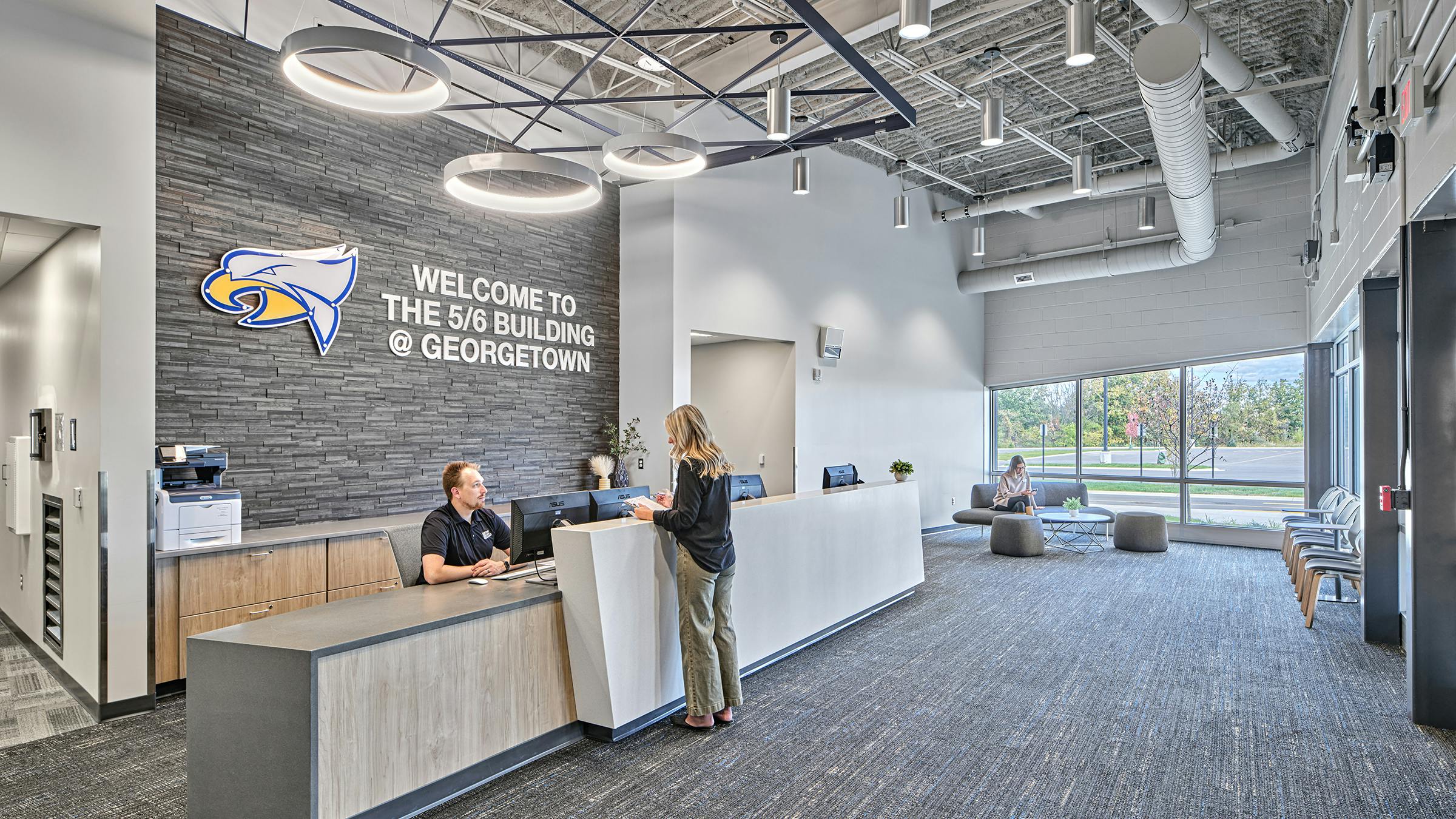 Male receptionist greets female visitor at reception desk with school logo against stone feature wall with circular lighting fixtures and exposed ductwork in the ceiling.