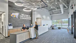 Male receptionist greets female visitor at reception desk with school logo against stone feature wall with circular lighting fixtures and exposed ductwork in the ceiling.